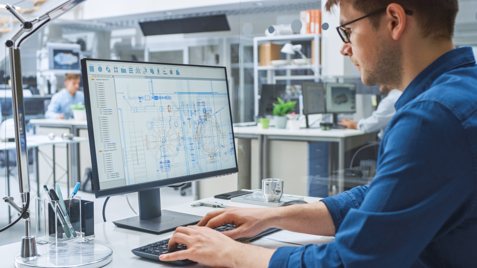 Over the Shoulder Shot of Engineer Working with CAD Software on Desktop Computer, Screen Shows Technical Drafts and Drawings. In the Background Engineering Facility Specialising on Industrial Design